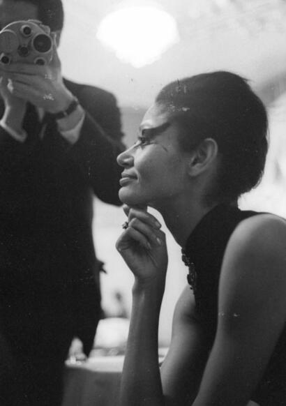 A woman with short hair and a choker necklace sits thoughtfully during a photoshoot, as a photographer captures her side profile.